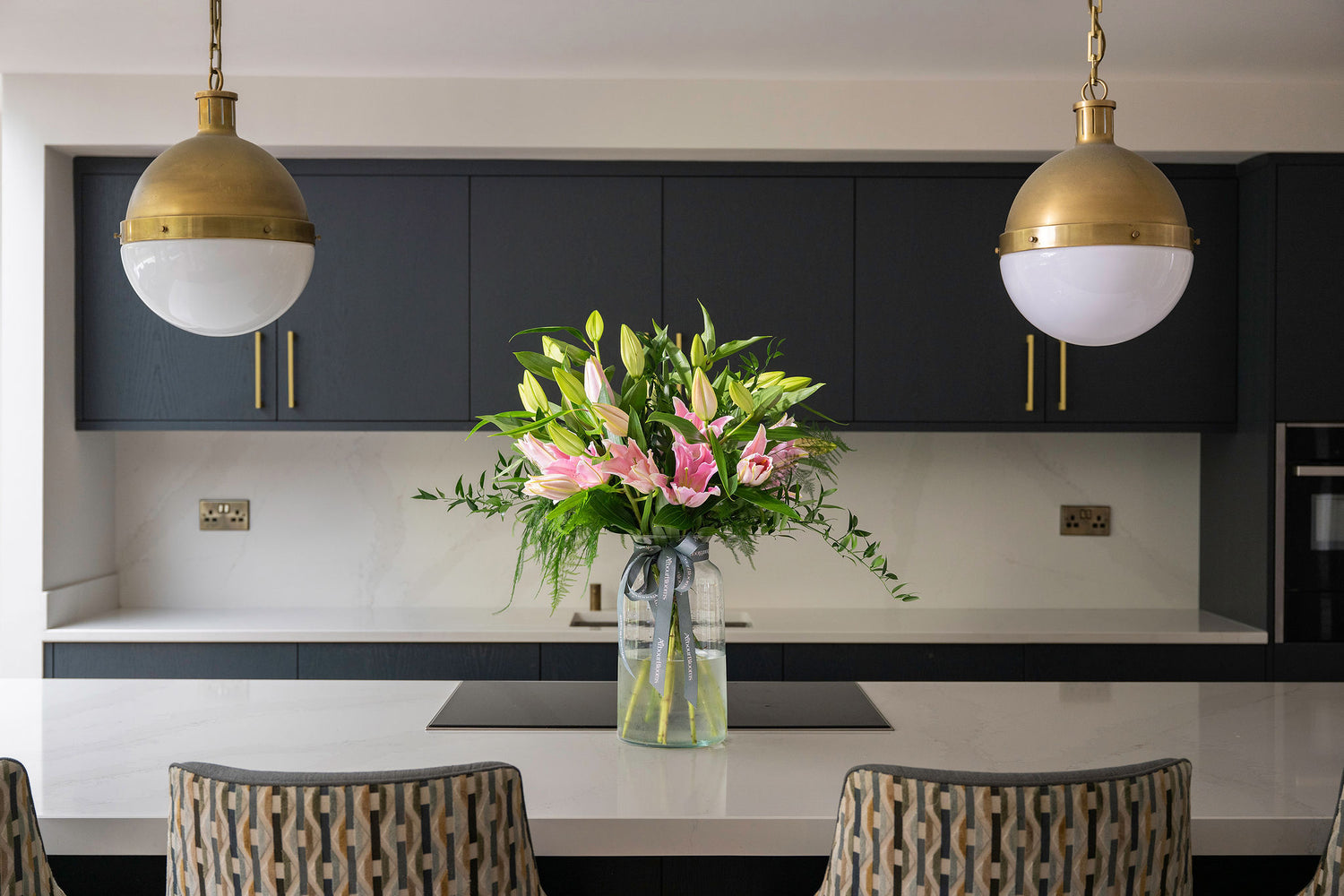 Vase of pink lilies on the counter of a modern kitchen with black cabinets and gold pendant lights