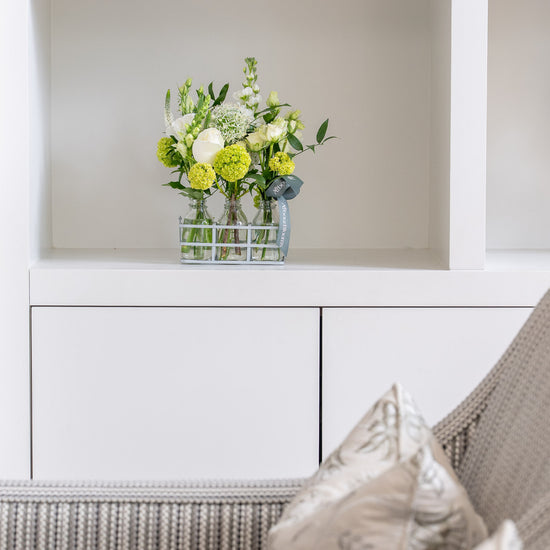 Floral arrangement in a glass vase on a white shelf above a wicker chair with cushions