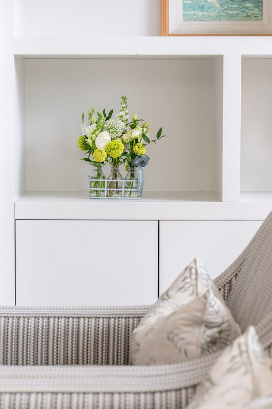 Floral arrangement in a glass vase on a white shelf above a wicker chair with cushions