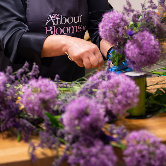 Florist arranging purple allium flowers on a wooden table, wearing an Arbour Blooms apron