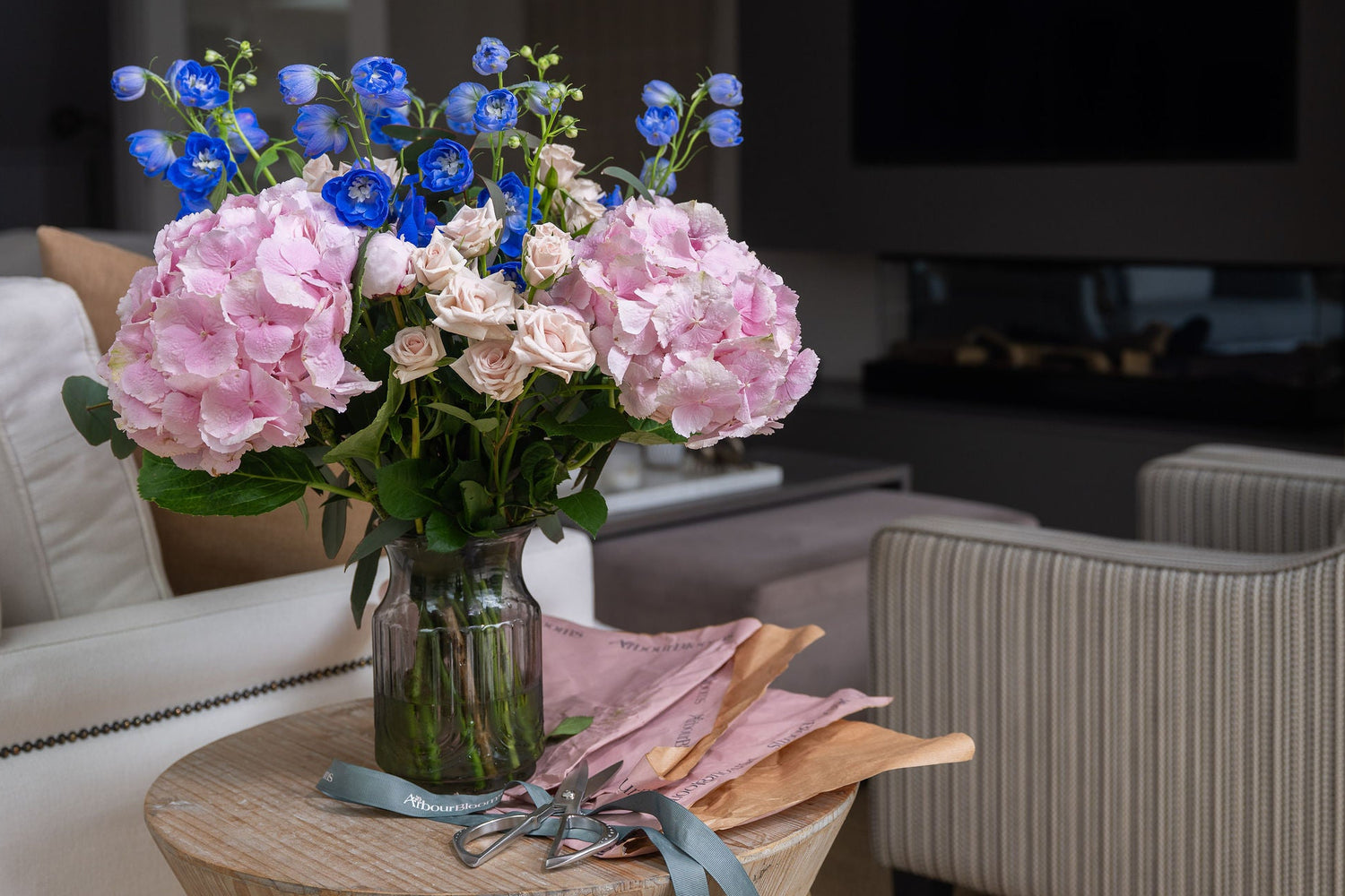 Bouquet of pink hydrangeas and blue delphiniums in a glass vase on a side table in a living room
