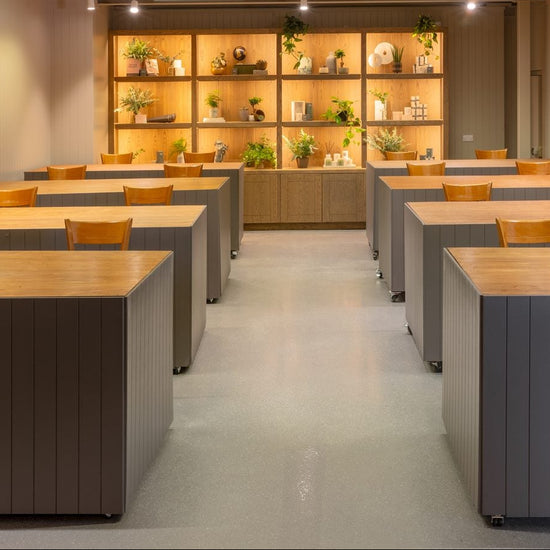 A modern indoor meeting or study space with multiple rectangular desks arranged in rows, featuring light wood tops and dark gray paneled bases. The room has beige walls, a central aisle, built-in shelving with warm lighting at the back, and ceiling-mounted track lights.