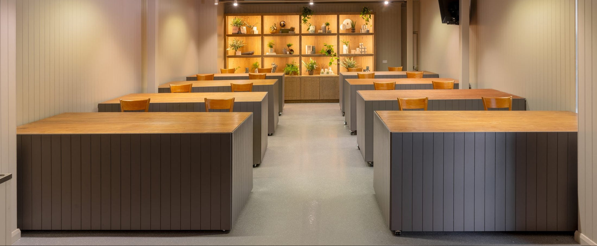 A modern indoor meeting or study space with multiple rectangular desks arranged in rows, featuring light wood tops and dark gray paneled bases. The room has beige walls, a central aisle, built-in shelving with warm lighting at the back, and ceiling-mounted track lights.